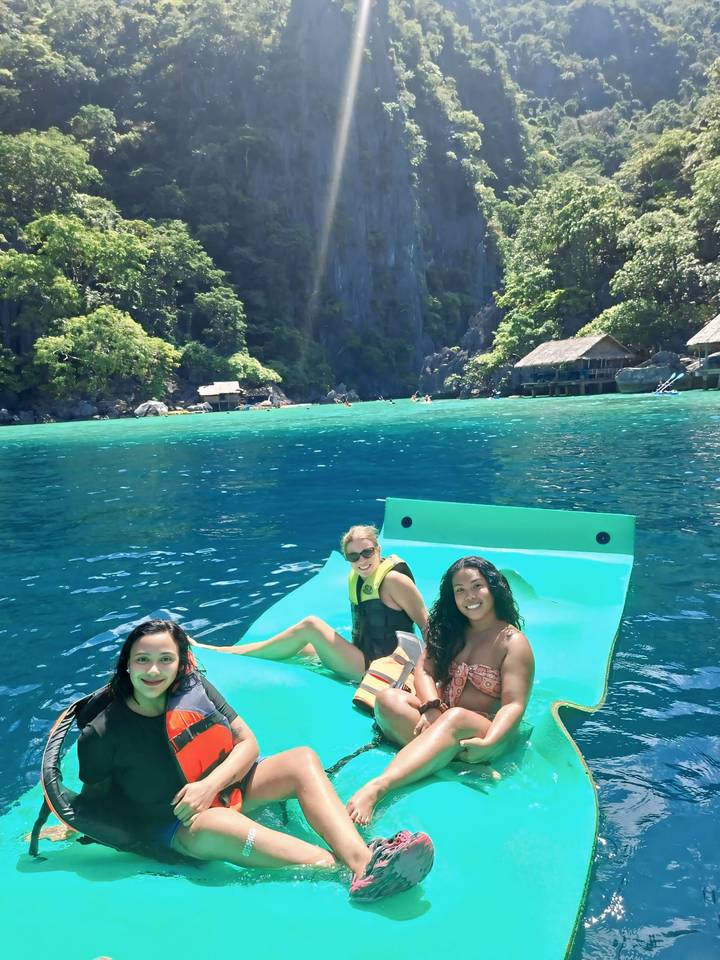 Three women lounge on a bright green floating mat in crystal-clear lagoon waters backed by towering limestone cliffs and jungle.