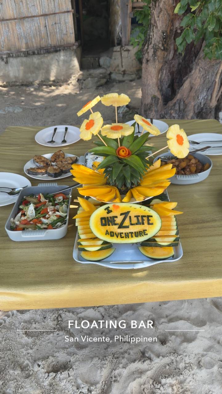 Colorful tropical fruit platter featuring a carved melon reading 'I ❤️ One Life Adventure' surrounded by sliced mango and salad dishes.