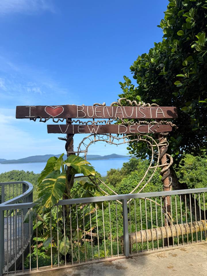 Rustic wooden sign reading 'I ❤ Buenavista View Deck' framed by vines with an island-studded bay in the background.