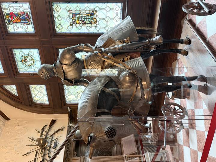 Museum display of an armored knight on horseback behind glass in a grand hall.