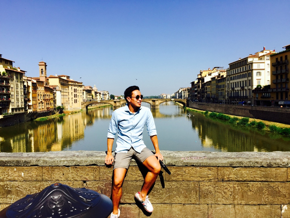 A person sitting on a stone wall overlooking a river and historic buildings.