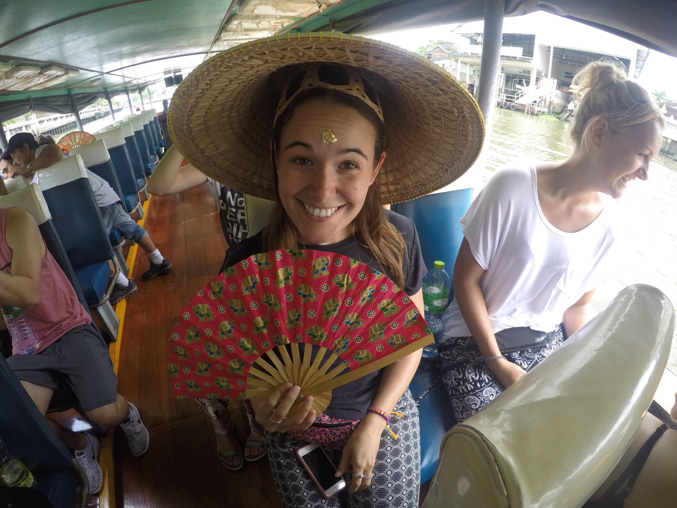 Woman on a boat, holding a fan and wearing a straw hat, smiling at camera.