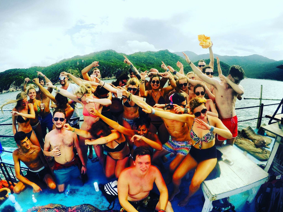 Large group of backpackers dabbing and celebrating on a party boat with jungle coastline behind.