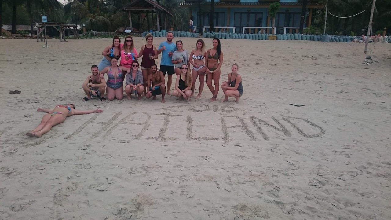 Group of people posing on a beach with 'THAILAND' written in the sand.