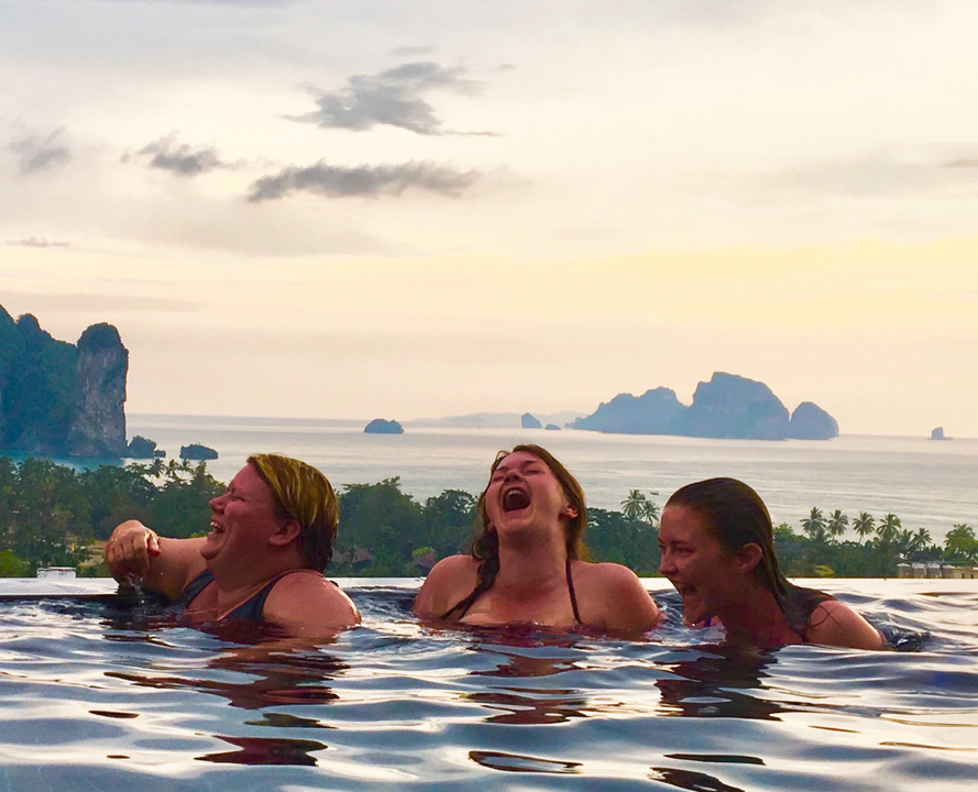 Three friends laughing in a pool with ocean and rock formation views.