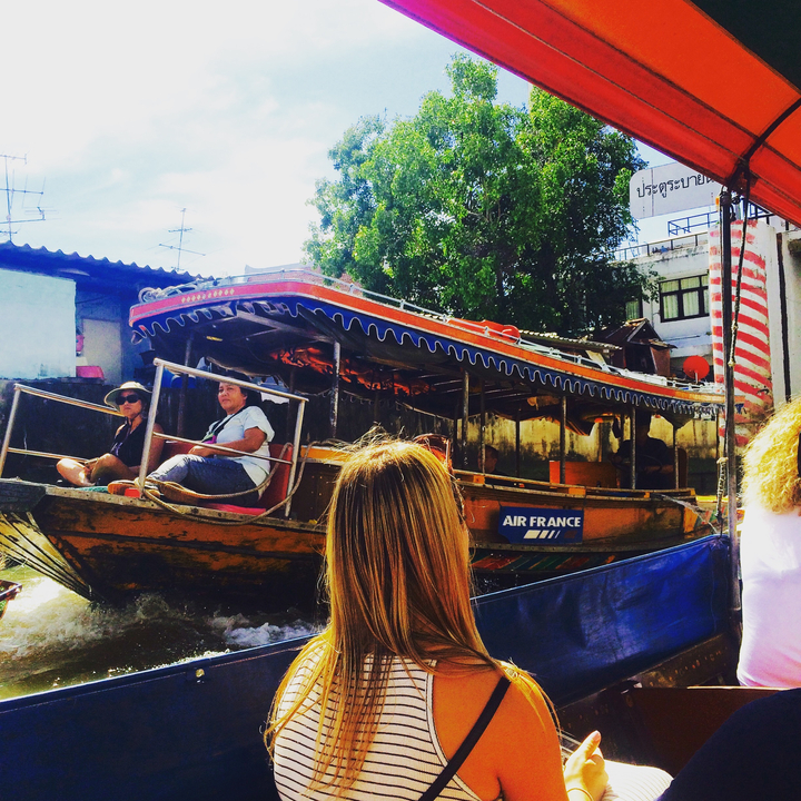 Thai longtail boat with people sitting, with vibrant decorations, on a canal.