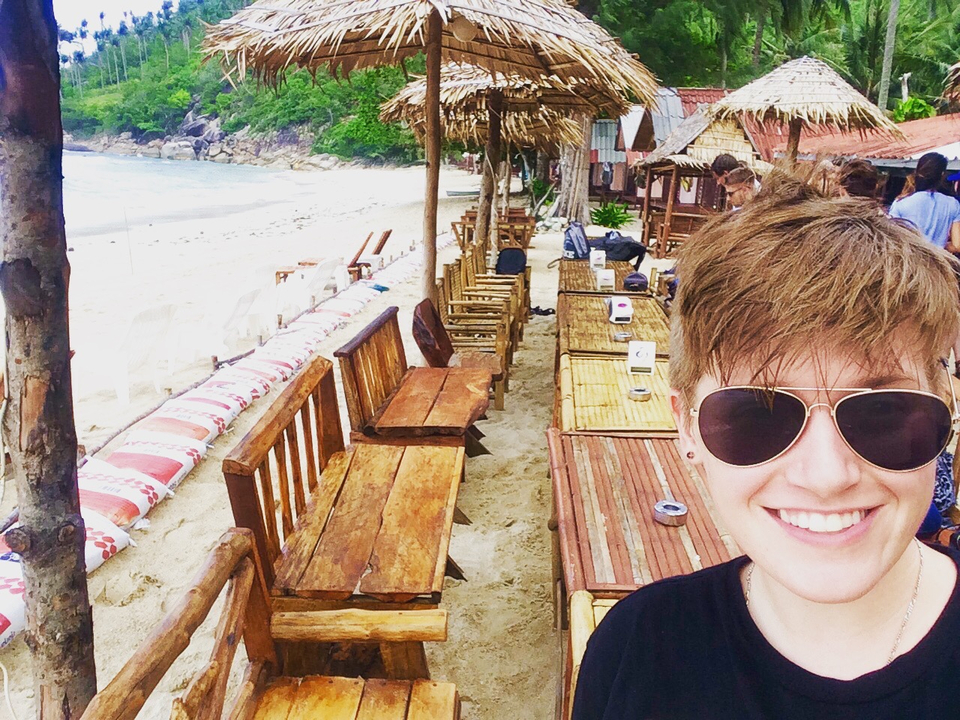 Person on a beach with tables and chairs under thatched umbrellas.