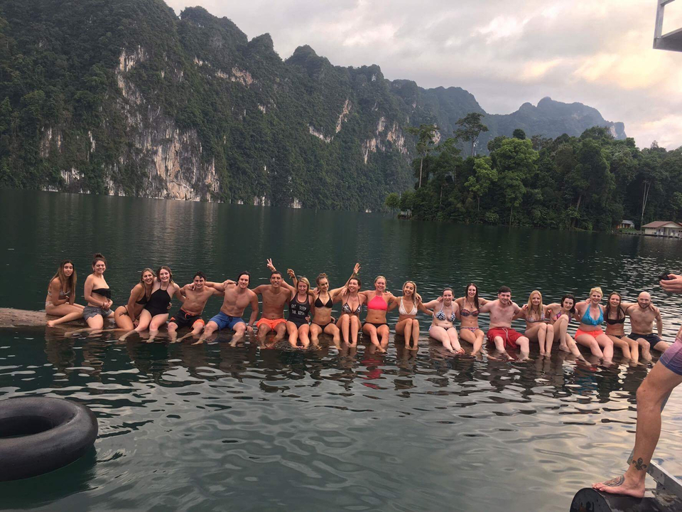 Group of people seated along a log in a lake with mountains.