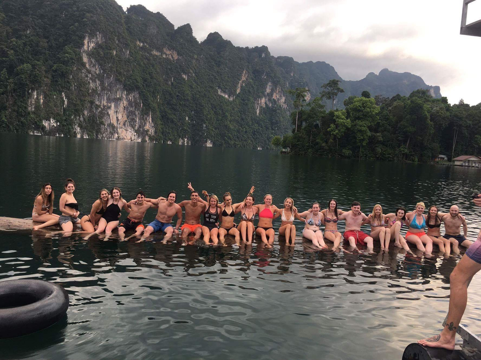 Group of people seated along a log in a lake with mountains.
