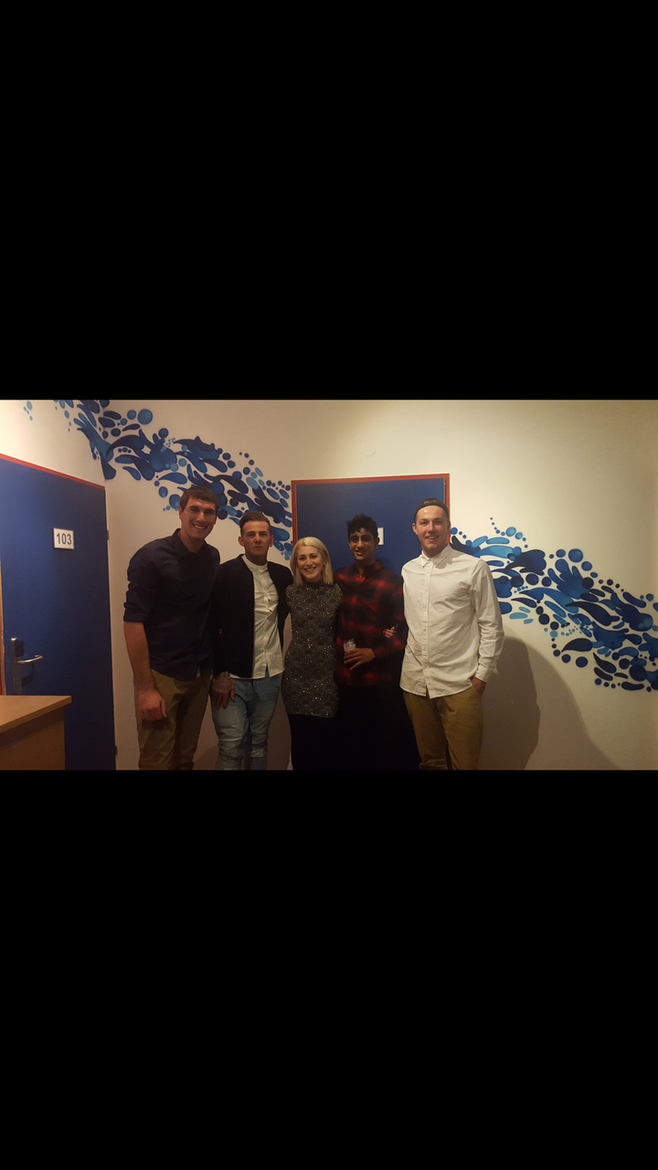 Group of people posing indoors with an artistic wall in the background.