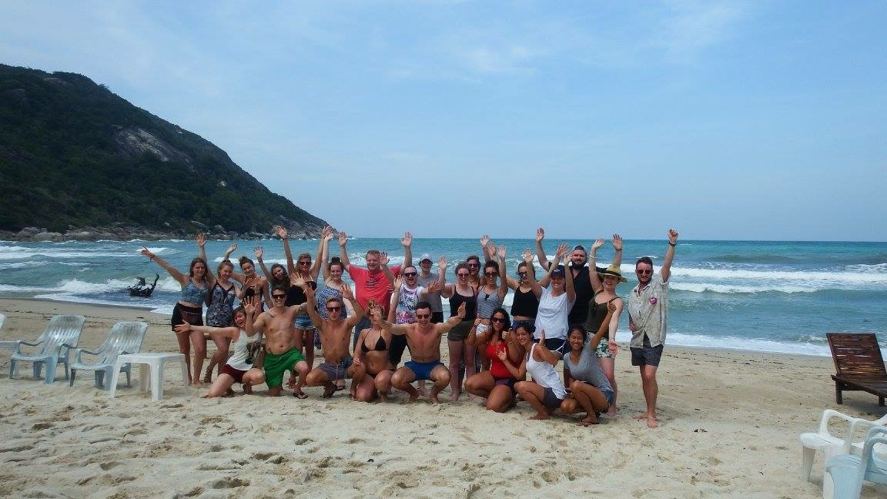 Large group of people posing on a beach with ocean and hills in the background.