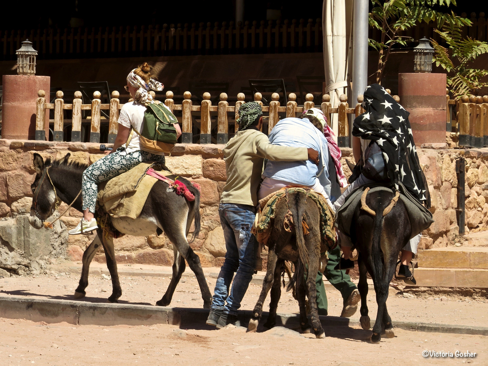 People riding donkeys, assisted by locals in a desert landscape.