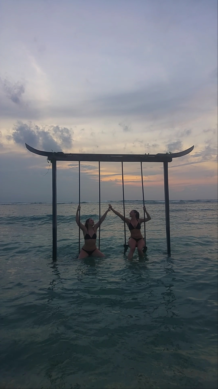 Two people on swing sets over the ocean during sunset