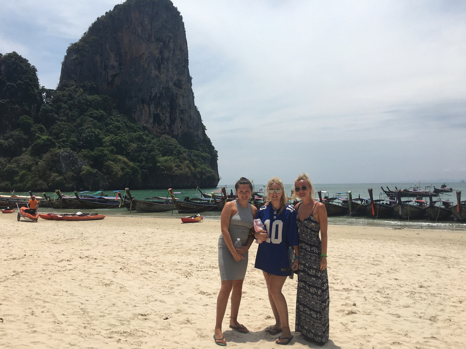 Three women standing on a beach with boats and a cliff in the background.