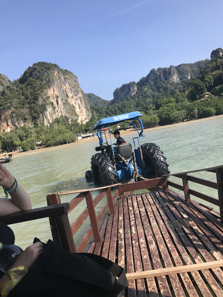 Man driving a tractor through shallow water with cliffs behind.