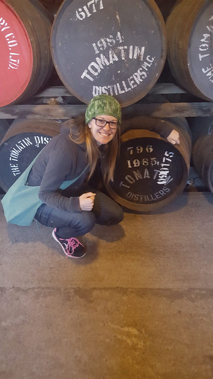 Person posing with whisky barrels at a distillery.