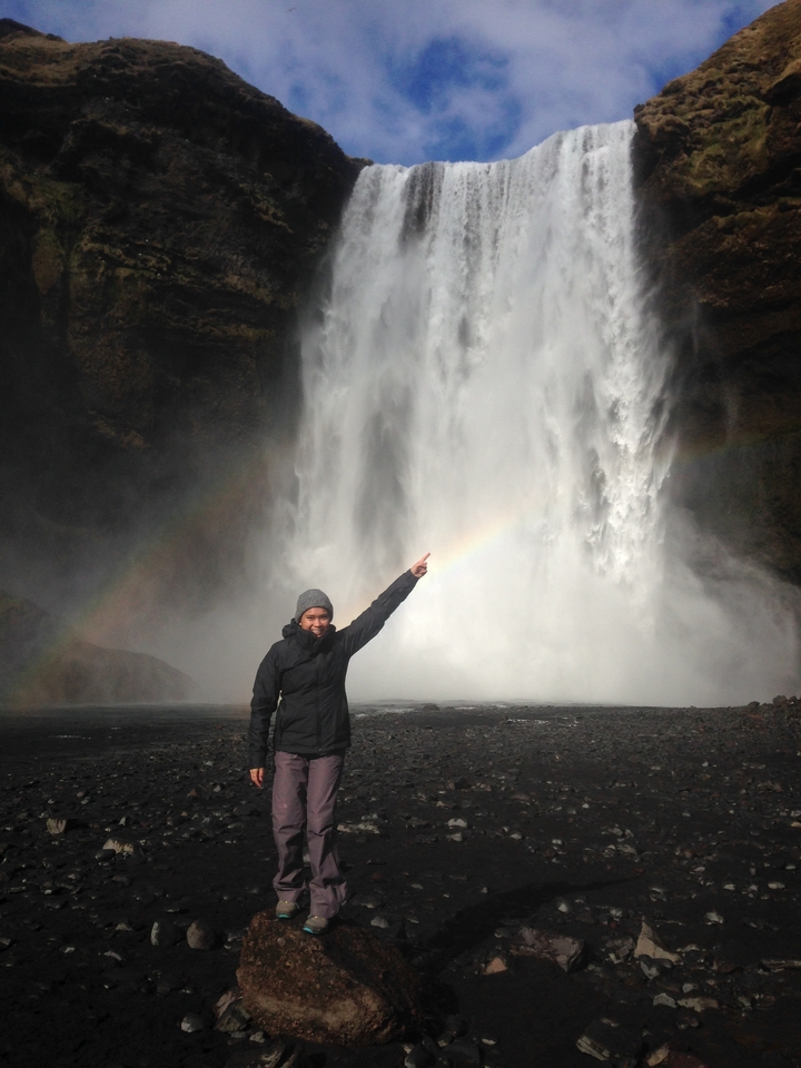 Person pointing at a waterfall with a rainbow in the mist.