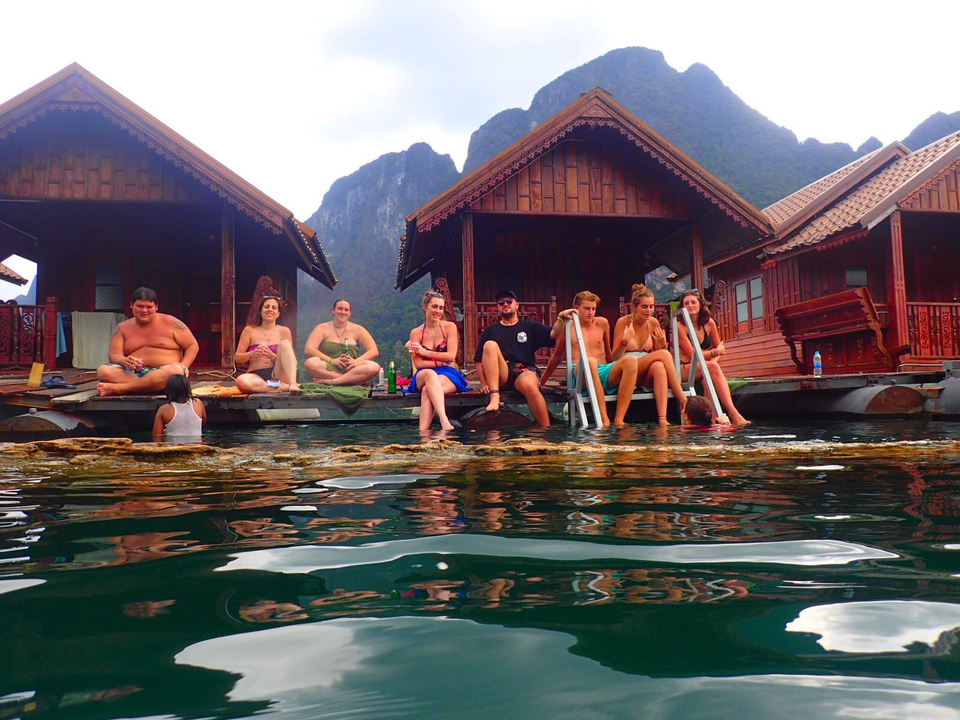 People sitting on the wooden planks by the water with lush mountains in the background.