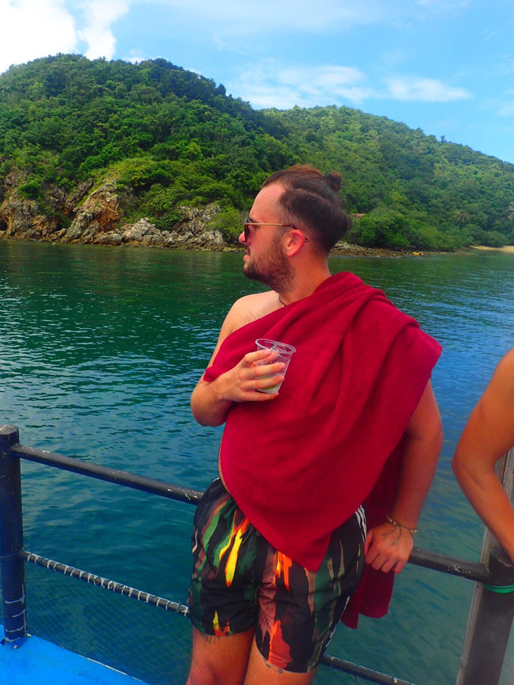 Person wrapped in a red towel standing by the water, holding a drink.