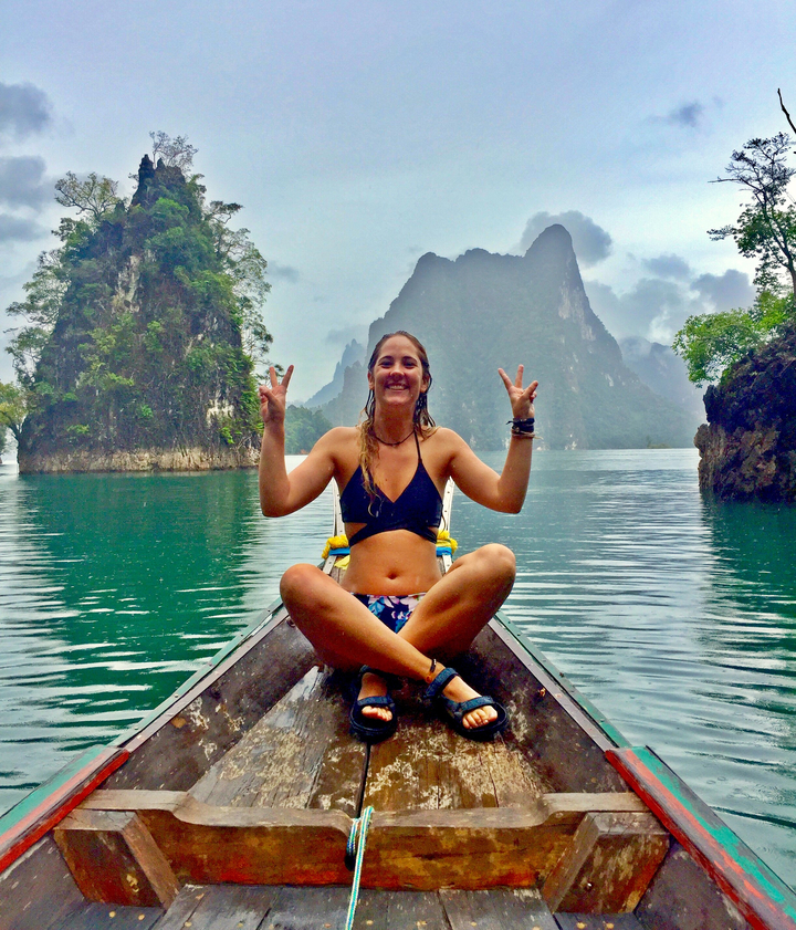 Person sitting on a boat making peace signs, with dramatic cliffs in the background.