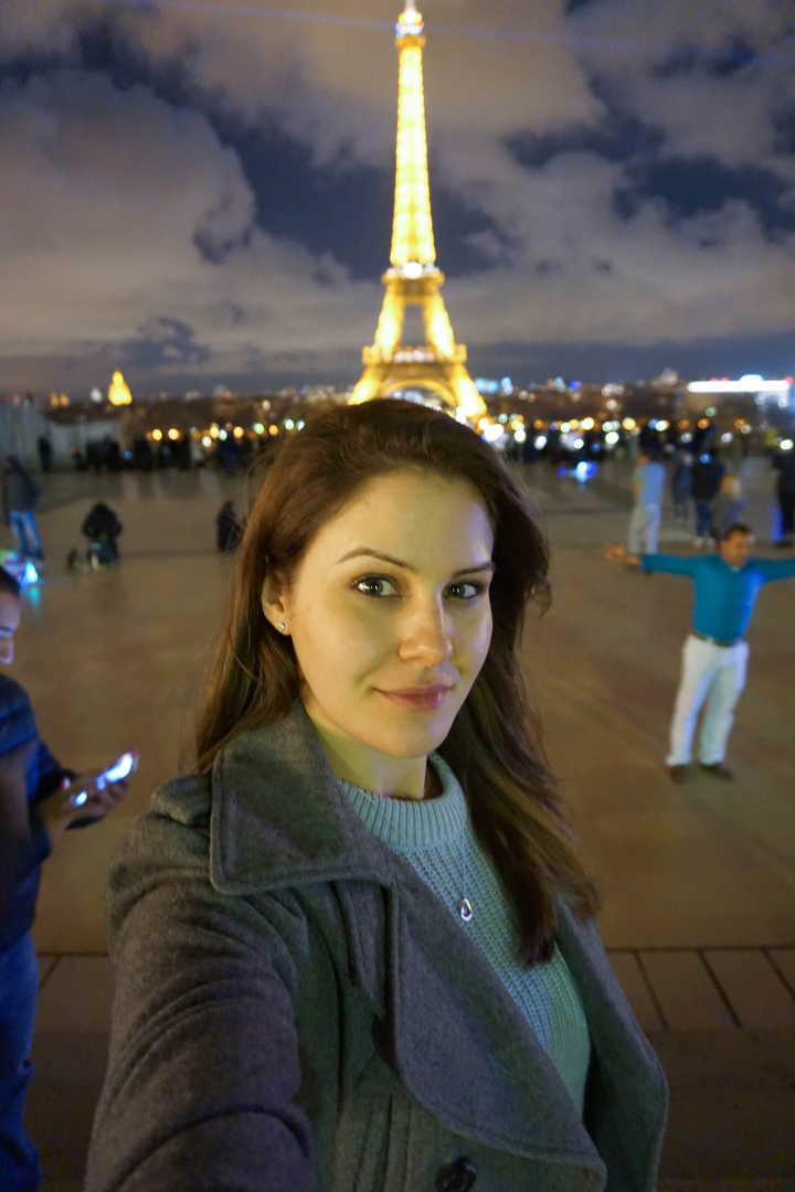 Person taking a selfie with a city scene in the evening.