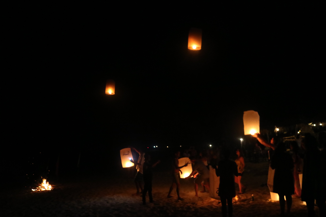 People releasing lanterns into the night sky by a bonfire.