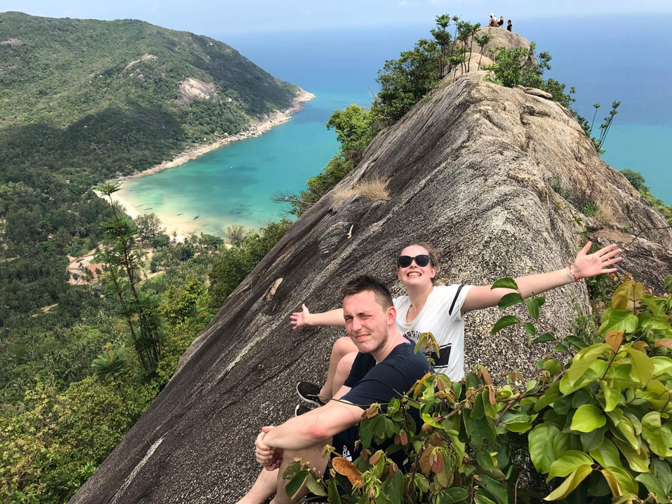 Two people on a cliff with a beach and blue water below.