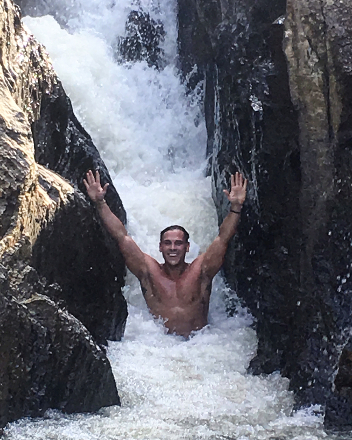 Person posing under a waterfall with arms raised.