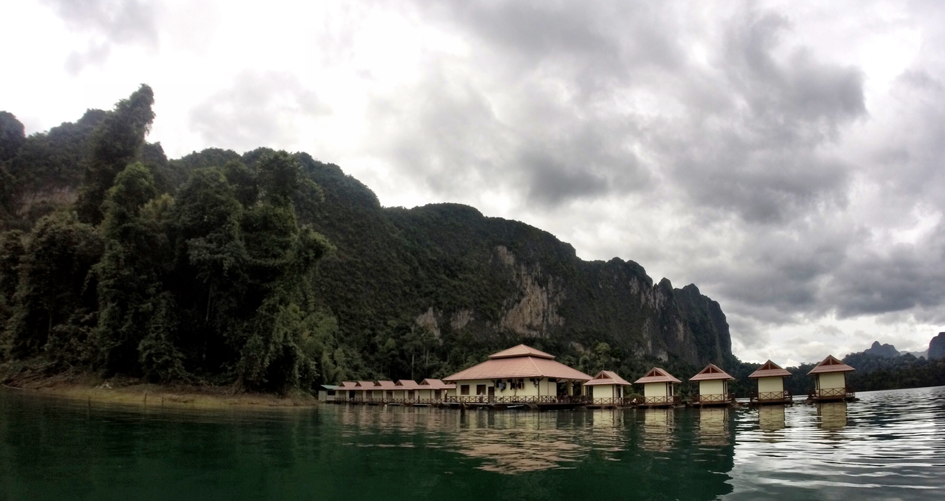 Floating bungalows on a lake surrounded by lush greenery under a cloudy sky.