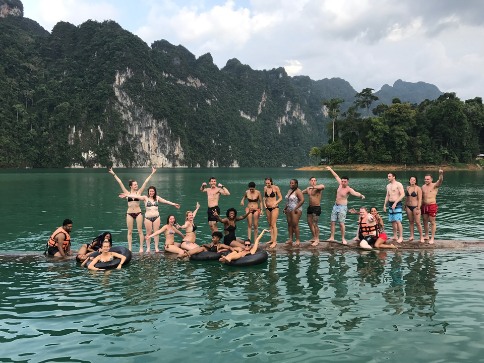 Group of people posing on a log in a lake with cliffs in the background.