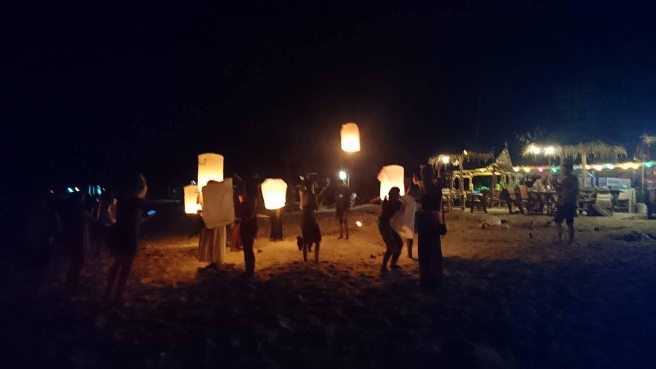 People releasing lanterns on a beach at night with lights in the background.