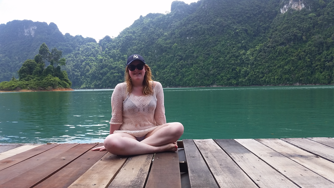 Person seated on a wooden deck by a serene lake in a hilly area.