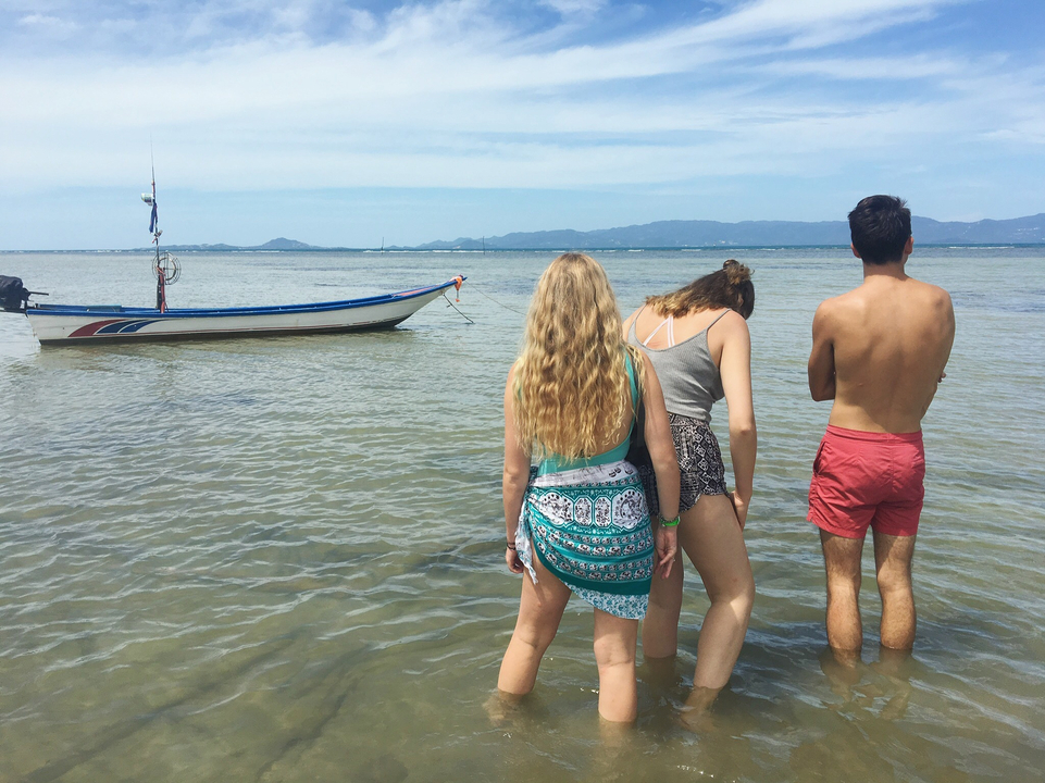Three people standing in shallow water on a beach with a boat nearby.