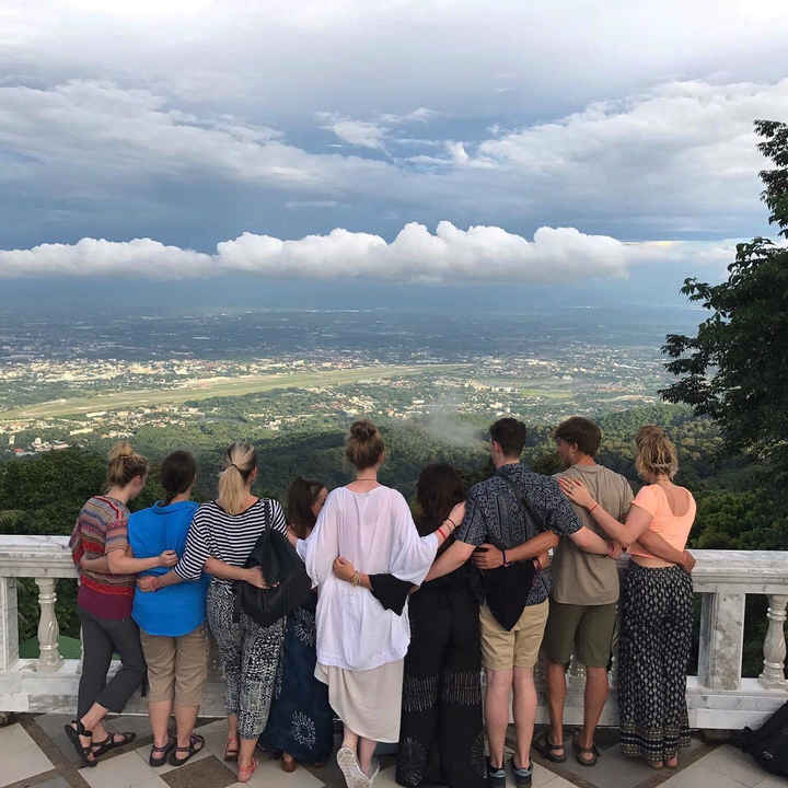 A group of people with their backs to the camera overlooking a scenic view from a mountain viewpoint.