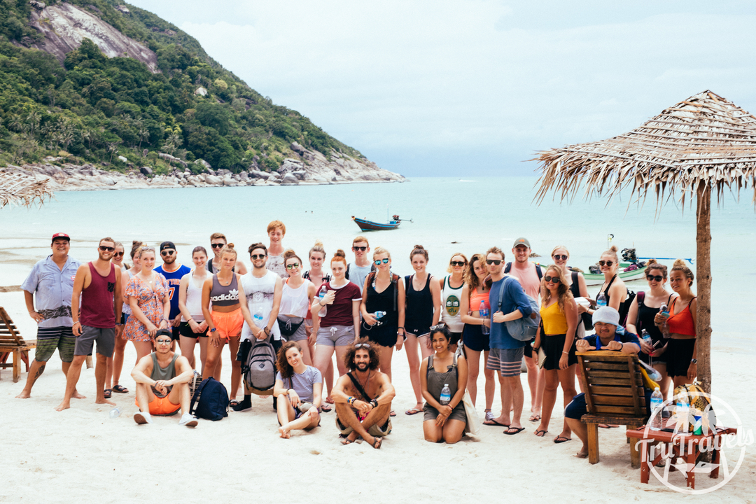 Group of people posing on a beach with a hut, watermark present.