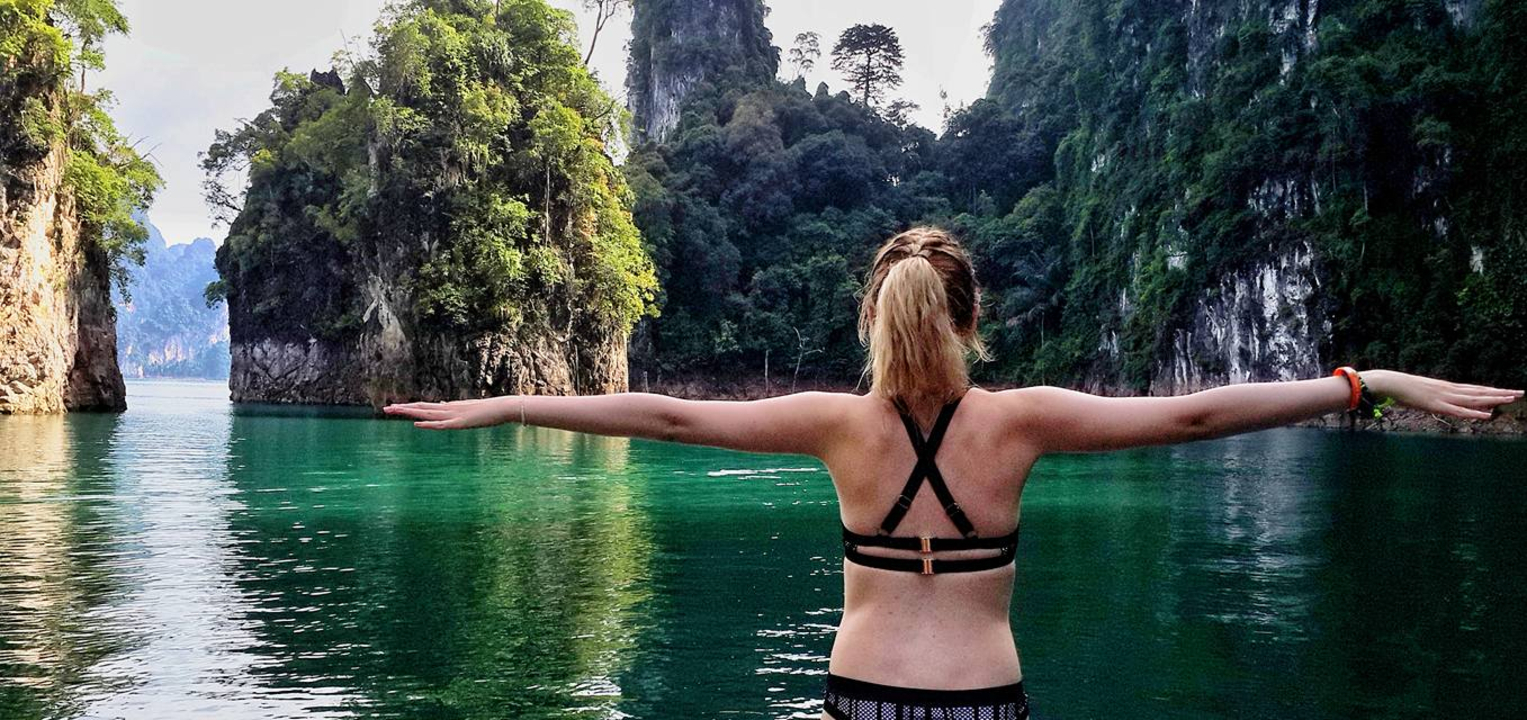 A person with arms outstretched in front of a picturesque lake and rock formations.