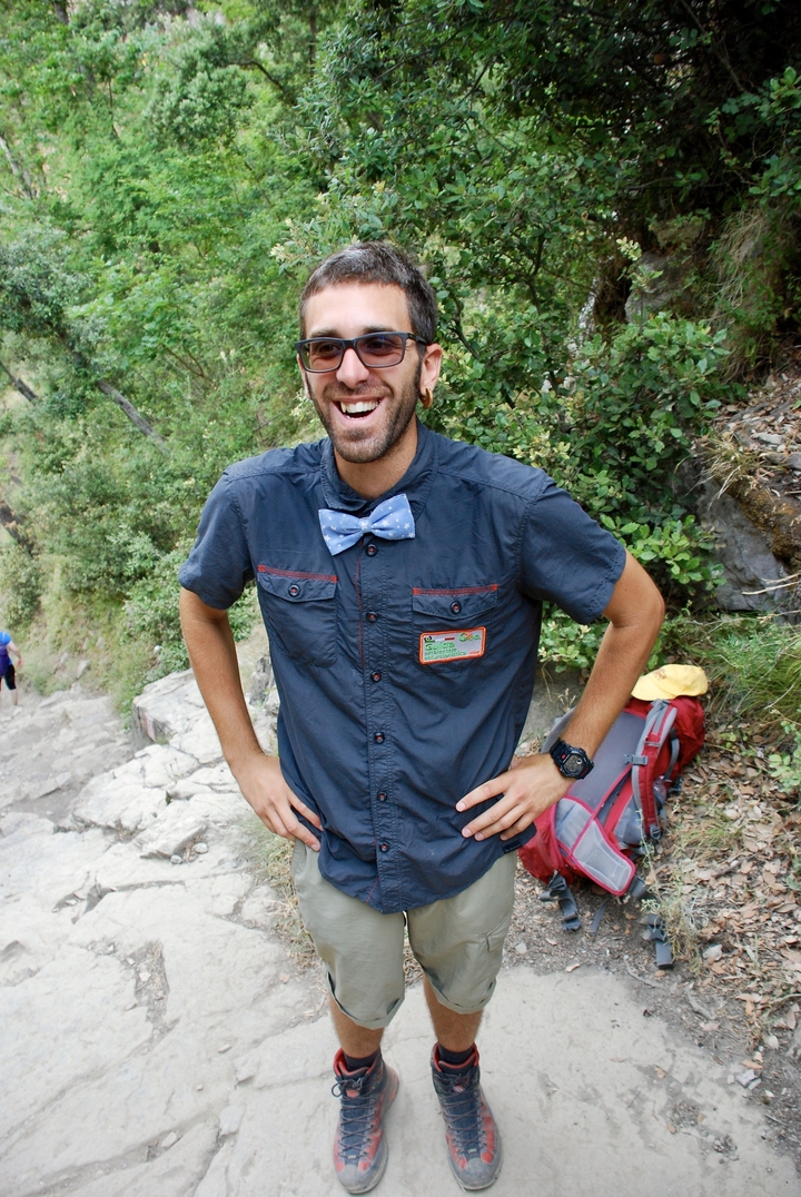 Homme souriant sur un sentier de randonnée, portant un nœud papillon et une casquette.
