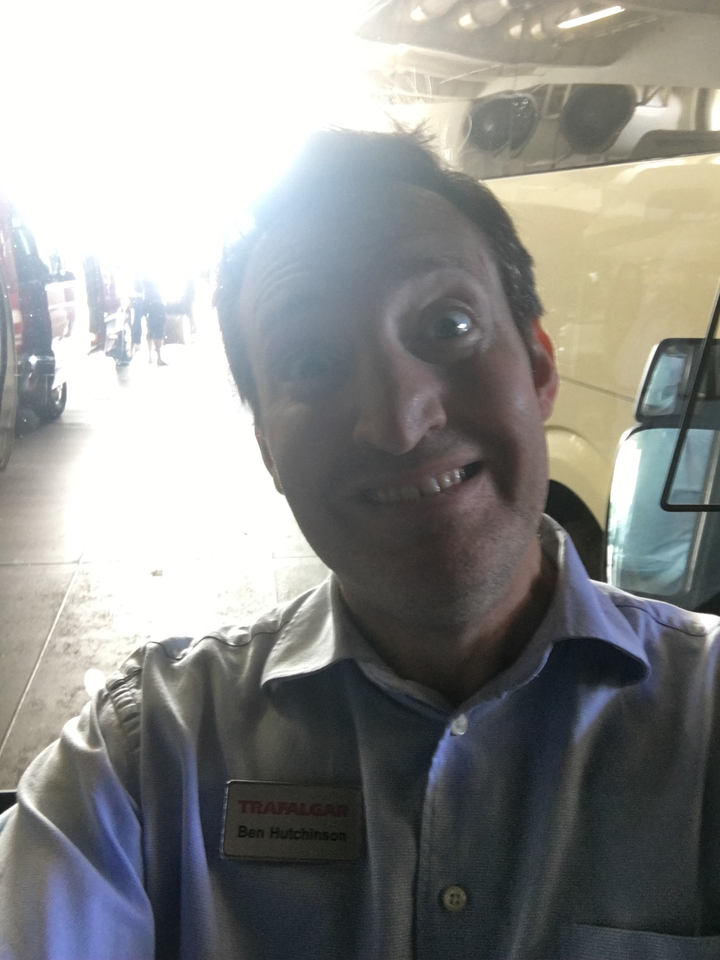 Gros plan d'une personne souriante dans une zone partiellement ombragée, avec des véhicules en arrière-plan.