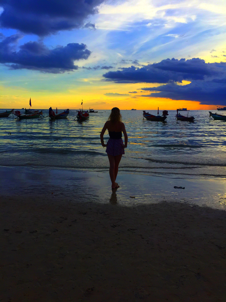 Silhouette of a person standing on a beach at sunset with fishing boats in the water.