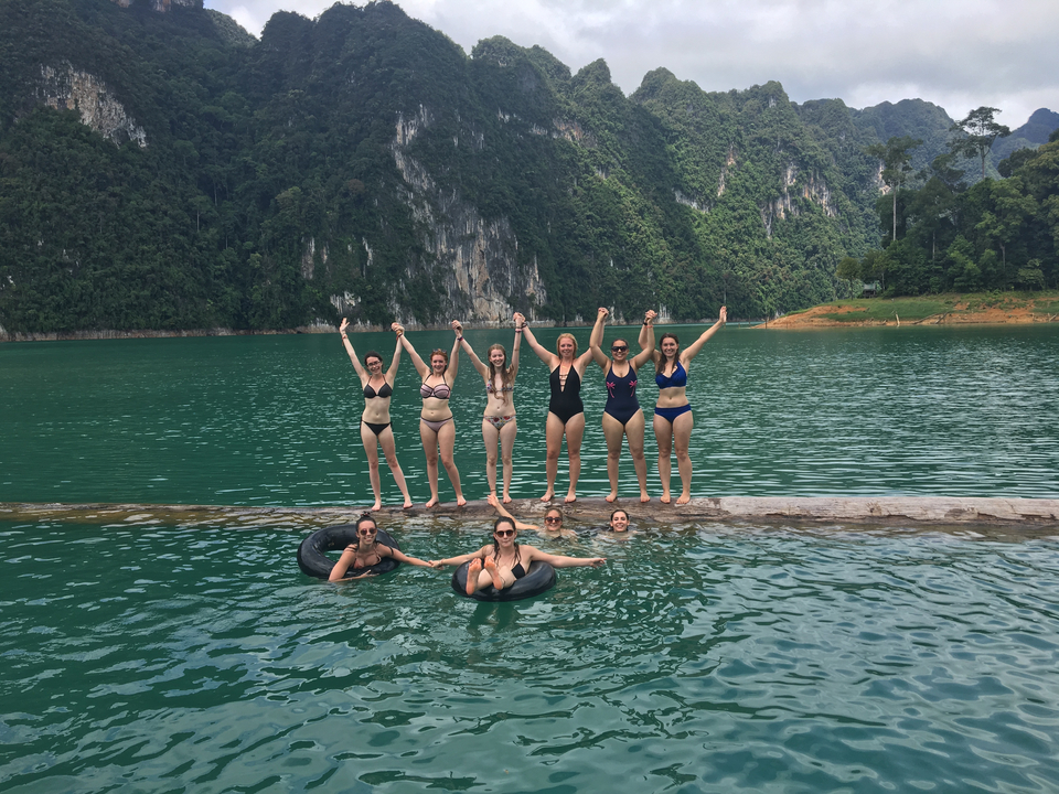 People in bikinis posing by a lake with cliffs and a forested backdrop.