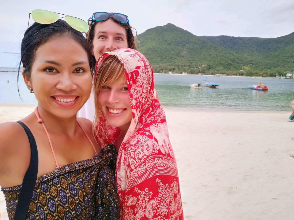 Smiling group on a beach with boats and hills in the background.