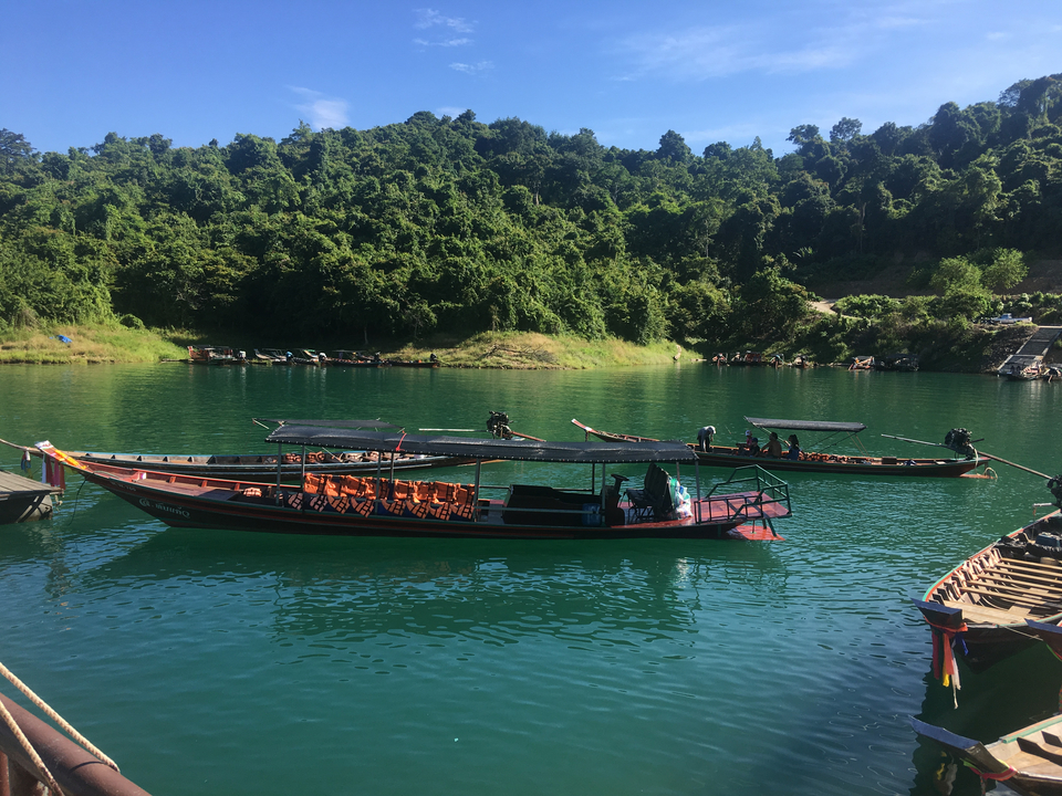 Longtail boats floating on emerald water against a backdrop of dense jungle hills under blue sky.