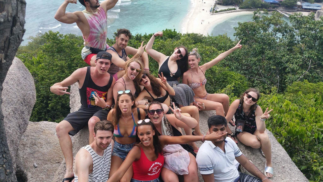 Large tour group cheerfully celebrating on a rocky viewpoint overlooking turquoise bays and green islands.