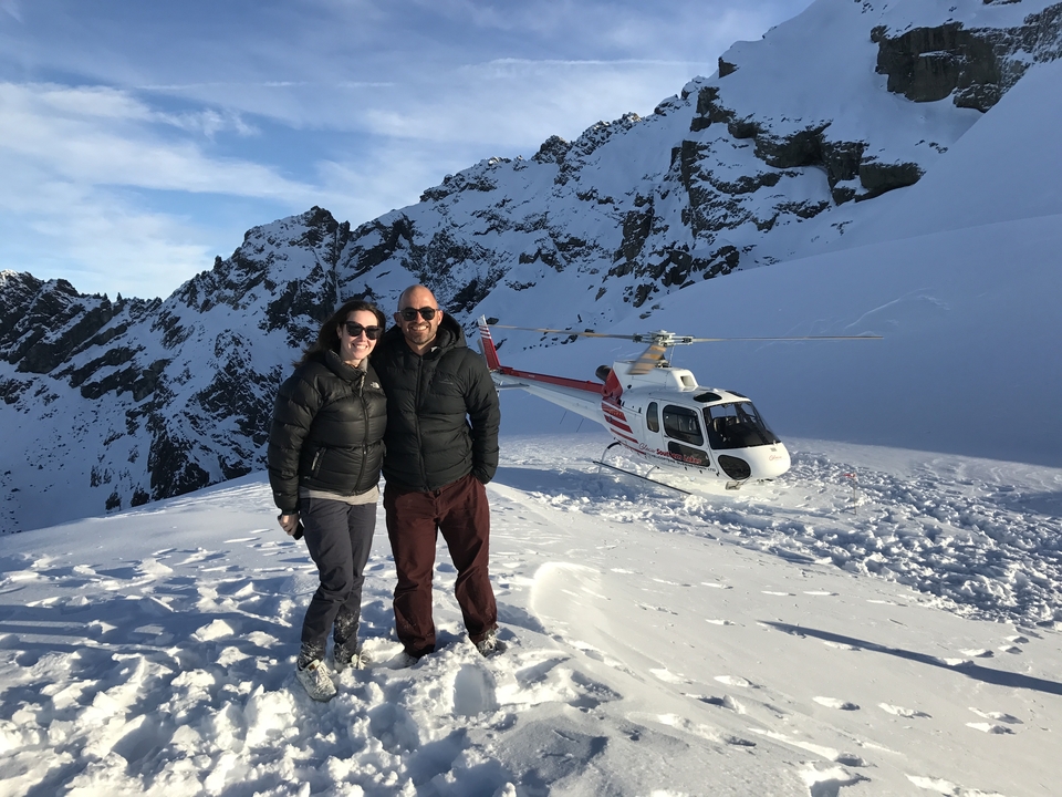 Couple standing on a snowy mountain with a helicopter in the background.