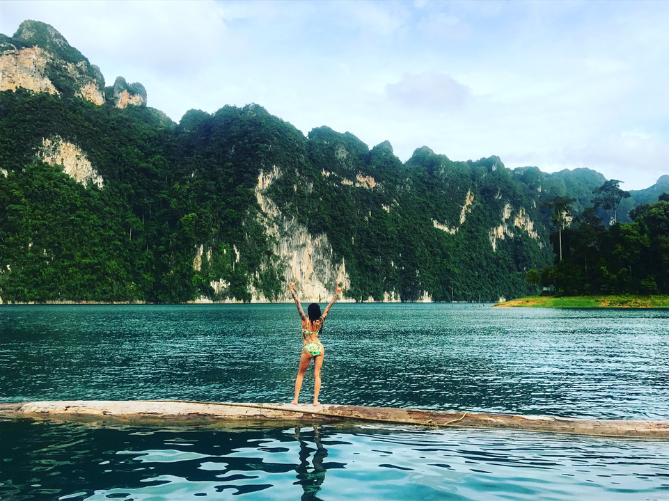 Person standing on a log facing a beautiful green mountain range.