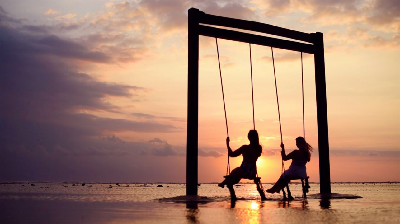 Silhouettes of people swinging over the ocean at sunset.