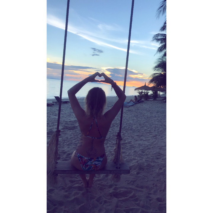 Person on a beach swing making a heart shape with hands during sunset.