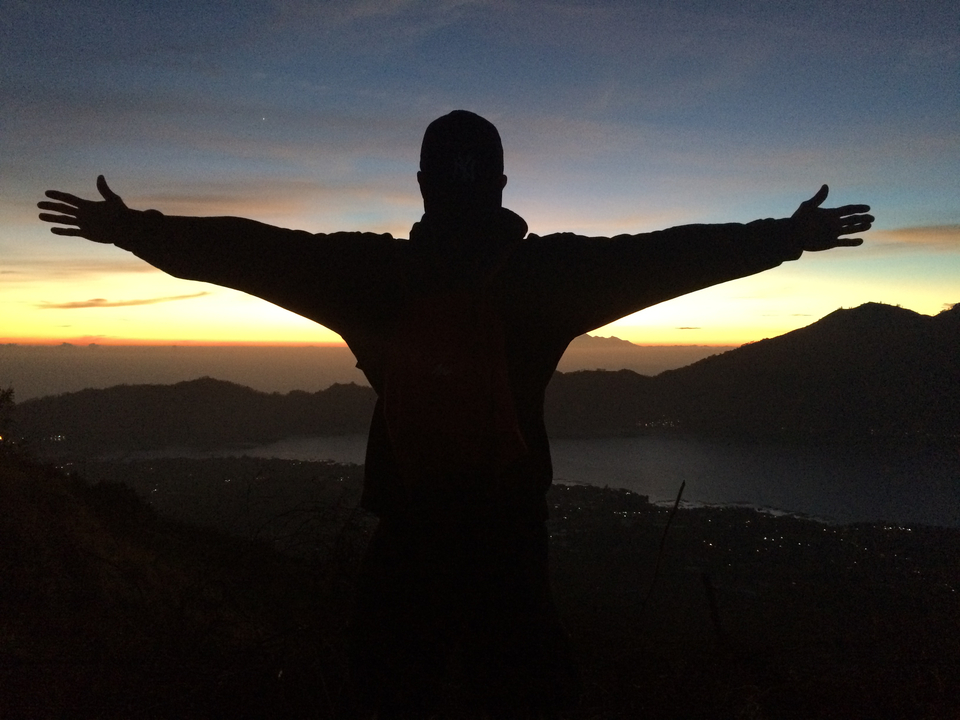 Silhouette of a person with arms outstretched on a mountain summit at dawn overlooking a lake and ridges.
