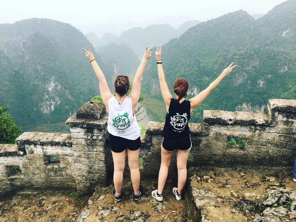 Two people with arms raised on a stone wall overlooking a scenic landscape.