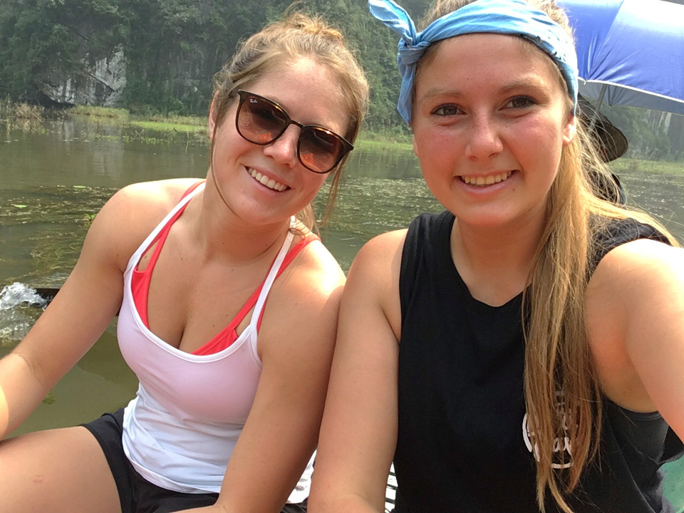 Two women smiling on a boat with greenery in the background.